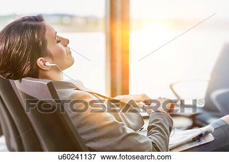 Stock Photo - Portrait of businesswoman listening to music on smartphone while sleeping and waiting for boarding in airport. Fotosearch