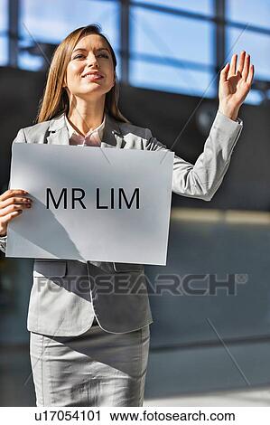 Stock Image - Portrait of businesswoman waving for her boss while holding blank white board in arrival area at airport. Fotosearch
