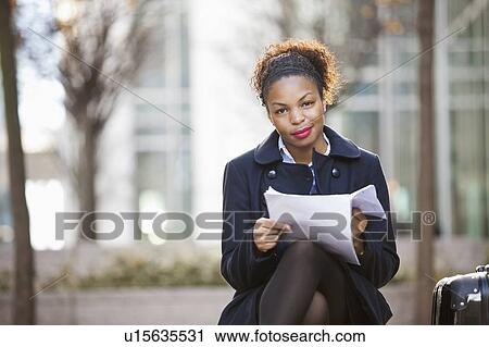 Stock Image - Portrait of confident African American young woman with documents sitting outdoors. Fotosearch