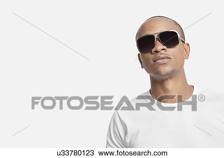 Stock Image - Portrait of confident young man wearing sunglasses against white background. Fotosearch