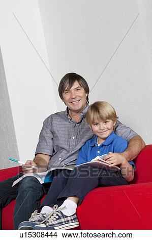 Picture - Portrait of father and son doing homework together while sitting on sofa. Fotosearch