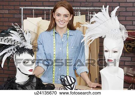Stock Image - Portrait of female fashion designer standing by table with fascinators on mannequin. Fotosearch