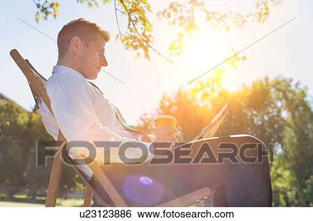 Stock Photograph - Portrait of handsome businessman drinking coffee and reading newspaper while sitting on folding chair in park. Fotosearch