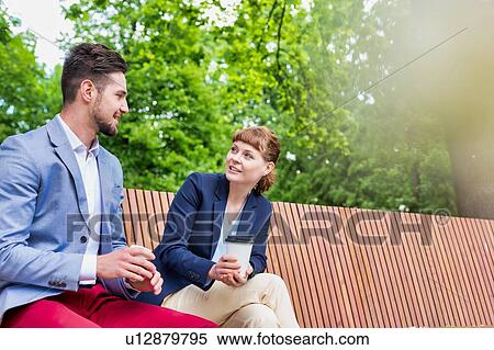 Stock Photography - Portrait of handsome businessman gesturing and talking to businesswoman while sitting on bench during office break. Fotosearch