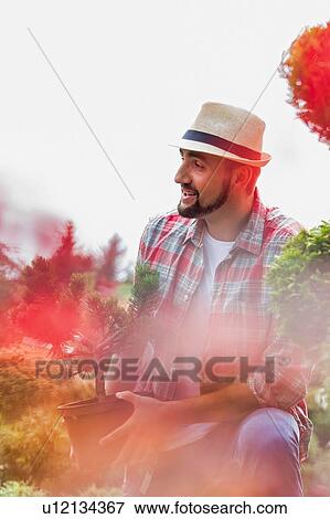 Stock Photo - Portrait of handsome gardener wearing hat while holding flower pot in the garden. Fotosearch