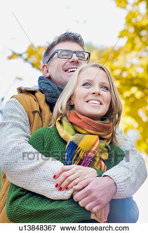 Stock Photo - Portrait of handsome man embracing his wife from the back in park. Fotosearch
