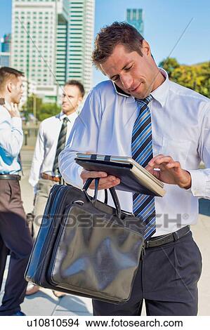 Picture - Portrait of handsome mature businessman talking on smartphone checking appointment on digital tablet. Fotosearch