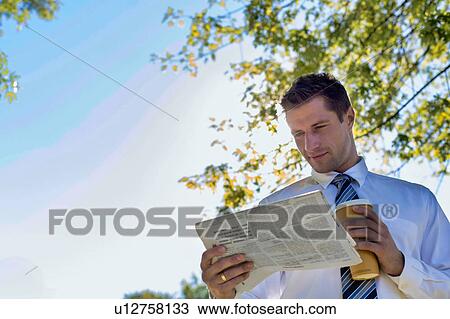 Stock Image - Portrait of handsome mature businessman holding cup of coffee while reading newspaper in park during break . Fotosearch