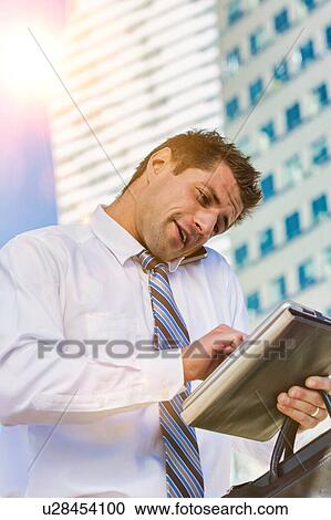 Stock Image - Portrait of handsome mature businessman talking on smartphone checking appointment on digital tablet. Fotosearch