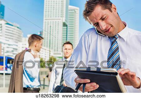 Stock Photo - Portrait of handsome mature businessman talking on smartphone checking appointment on digital tablet. Fotosearch