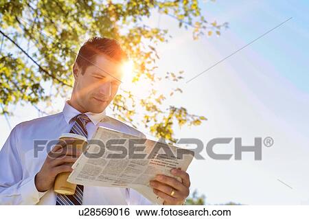 Stock Photograph - Portrait of handsome mature businessman holding cup of coffee while reading newspaper in park during break . Fotosearch