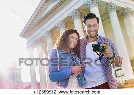 Stock Image - Portrait of handsome young man showing pictures on camera to his beautiful girlfriend while standing against museum with lens flare in background. Fotosearch
