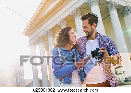 Stock Image - Portrait of handsome young man showing pictures on camera to his beautiful girlfriend while standing against museum with lens flare in background. Fotosearch