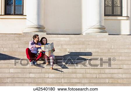 Stock Image - Portrait of handsome young man showing pictures on camera to his beautiful girlfriend while sitting against museum with lens flare in background. Fotosearch