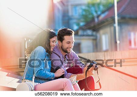 Stock Photo - Portrait of handsome young man showing pictures on camera to his beautiful girlfriend while sitting against museum with lens flare in background. Fotosearch