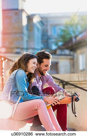 Stock Photograph - Portrait of handsome young man showing pictures on camera to his beautiful girlfriend while sitting against museum with lens flare in background. Fotosearch