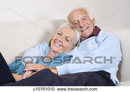 Stock Image - Portrait of happy elderly man with arm around spouse lying on sofa at home. Fotosearch