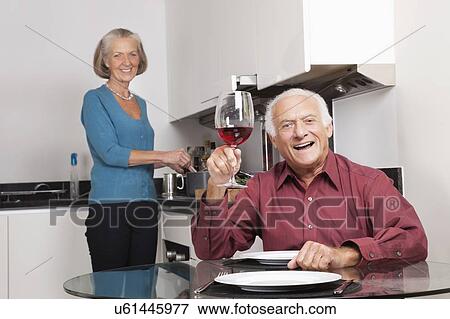Stock Photo - Portrait of happy senior couple at kitchen while man holding up wine glass. Fotosearch