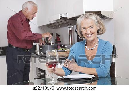 Stock Photo - Portrait of happy senior woman with wine glass while husband cooking food in kitchen. Fotosearch