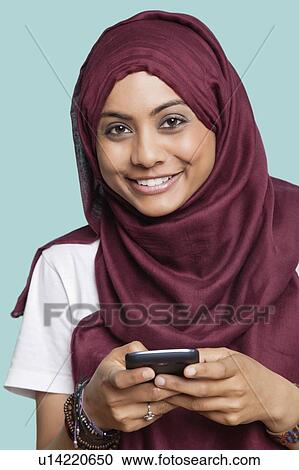 Stock Image - Portrait of happy young Muslim woman using cell phone against blue background. Fotosearch