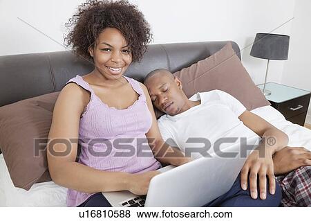 Stock Photo - Portrait of happy young woman using laptop while men sleeping in bed. Fotosearch