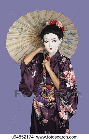 Picture - Portrait of Japanese woman in kimono with painted face holding parasol against purple background. Fotosearch
