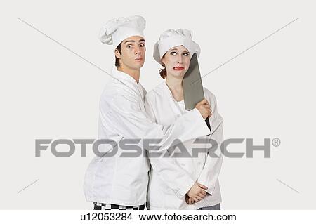 Picture - Portrait of male chef attacking female colleague with a cleaver against gray background. Fotosearch
