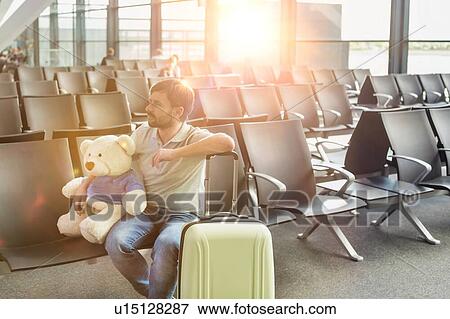 Stock Photo - Portrait of man sitting with teddy bear and suit case while waiting for boarding in airport. Fotosearch