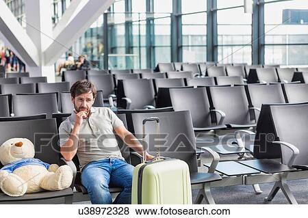 Stock Photo - Portrait of man sitting with teddy bear and suit case while waiting for boarding in airport. Fotosearch