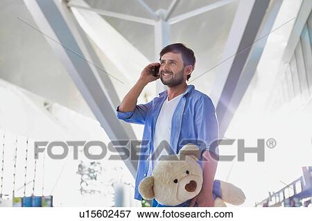 Stock Photo - Portrait of man talking on smartphone while holding a teddy bear present for his daughter in airport. Fotosearch