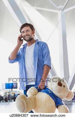 Stock Photo - Portrait of man talking on smartphone while holding a teddy bear present for his daughter in airport. Fotosearch
