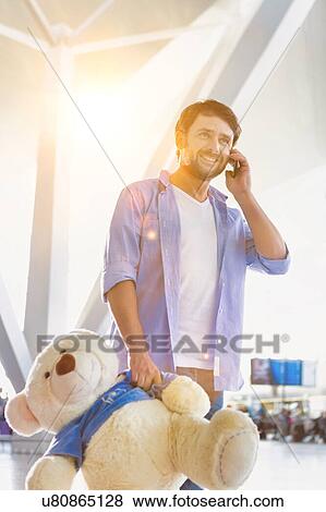 Stock Photo - Portrait of man talking on smartphone while holding a teddy bear present for his daughter in airport. Fotosearch