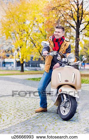 Stock Image - Portrait of mature attractive man sitting on motor bike while waiting in park. Fotosearch