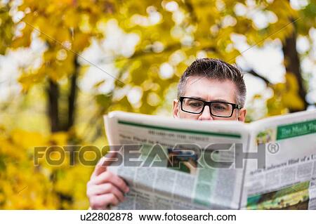 Stock Photo - Portrait of mature attractive man sitting on bench while reading newspaper in park. Fotosearch