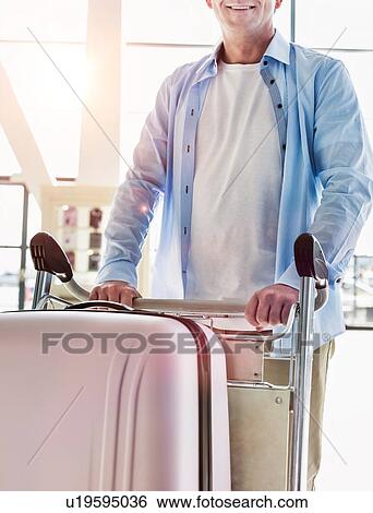 Stock Photograph - Portrait of mature attractive man pushing cart with his suitcase for check in at airport. Fotosearch