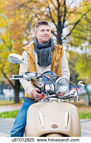 Stock Photography - Portrait of mature attractive man sitting on motor bike while waiting in park. Fotosearch