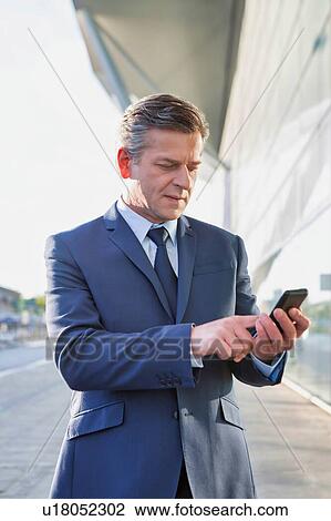 Stock Image - Portrait of mature attractive businessman using smartphone while standing in front of airport. Fotosearch