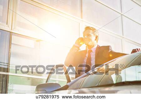 Stock Photography - Portrait of mature attractive businessman talking on smartphone while standing outside the car. Fotosearch