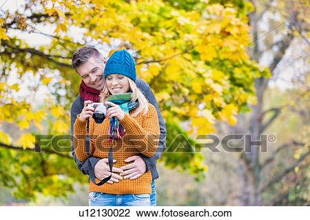 Stock Image - Portrait of mature beautiful woman showing pictures on camera while her husband is hugging her from behind in park. Fotosearch
