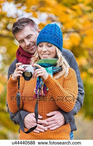 Stock Photo - Portrait of mature beautiful woman showing pictures on camera while her husband is hugging her from behind in park. Fotosearch