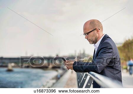Stock Photograph - Portrait of mature businessman leaning on railing while using smartphone. Fotosearch