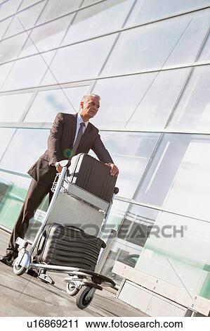 Stock Image - Portrait of mature businessman pushing luggage cart with his suitcase in airport. Fotosearch