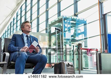 Stock Image - Portrait of Mature businessman sitting and holding his passport while waiting in gate for boarding at airport. Fotosearch