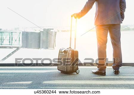 Picture - Portrait of mature businessman standing with his suitcase while looking through the window in airport. Fotosearch