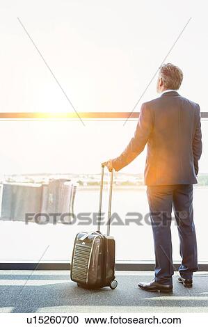 Stock Image - Portrait of mature businessman standing with his suitcase while looking through the window in airport. Fotosearch