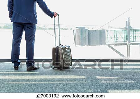 Stock Image - Portrait of mature businessman standing with his suitcase while looking through the window in airport. Fotosearch