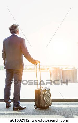 Stock Image - Portrait of mature businessman standing with his suitcase while looking through the window in airport. Fotosearch