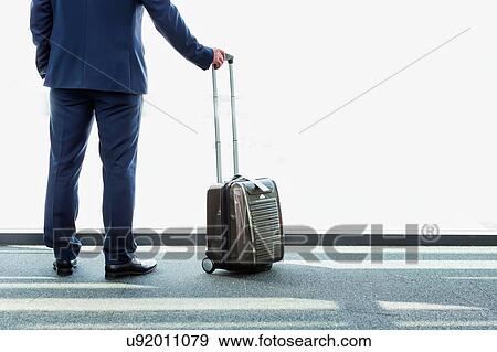 Stock Photo - Portrait of mature businessman standing with his suitcase while looking through the window in airport. Fotosearch