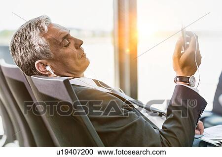 Portrait of mature businessman listening to music on his smartphone while waiting for boarding in airport View Large Photo Image Stock Image - Portrait of mature businessman listening to music on his smartphone while waiting for boarding in airport. Fotosearch