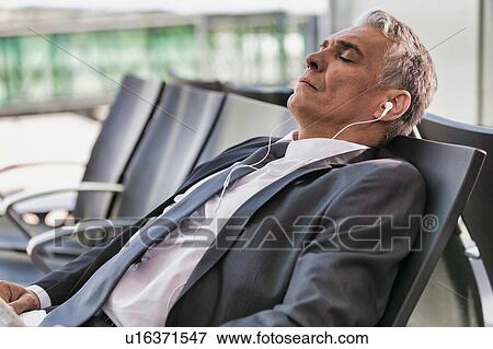 Stock Photo - Portrait of mature businessman listening to music on his smartphone while sleeping and waiting for boarding in airport. Fotosearch
